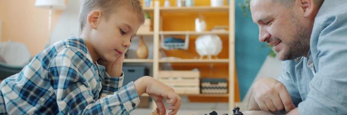 Smart child playing chess with dad at home lying on floor together having fun with intellectual sports game. Happy family and leisure activity concept.